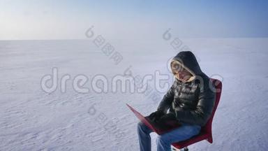 穿着保暖外套和头罩的商人坐在雪地里，手提电脑，双手放在头上，看着太阳